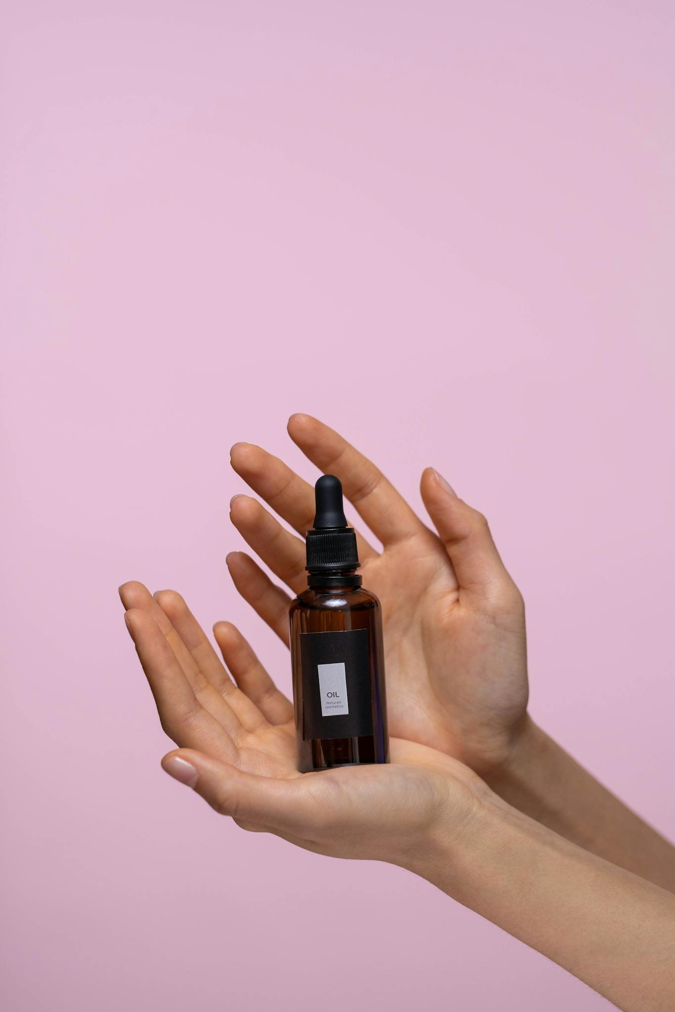 Close-up of a brown dropper bottle held by hands against a pink background, representing natural skincare.