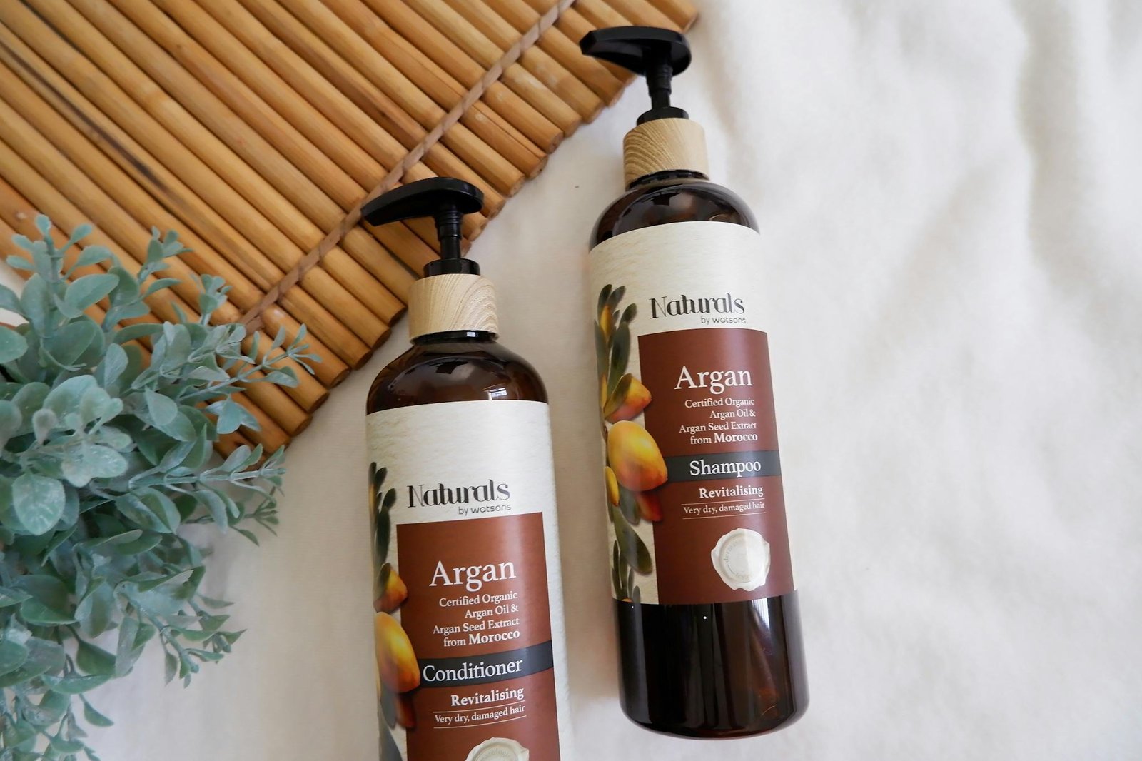 Close-up of argan oil shampoo and conditioner bottles with leaves on a bamboo mat.